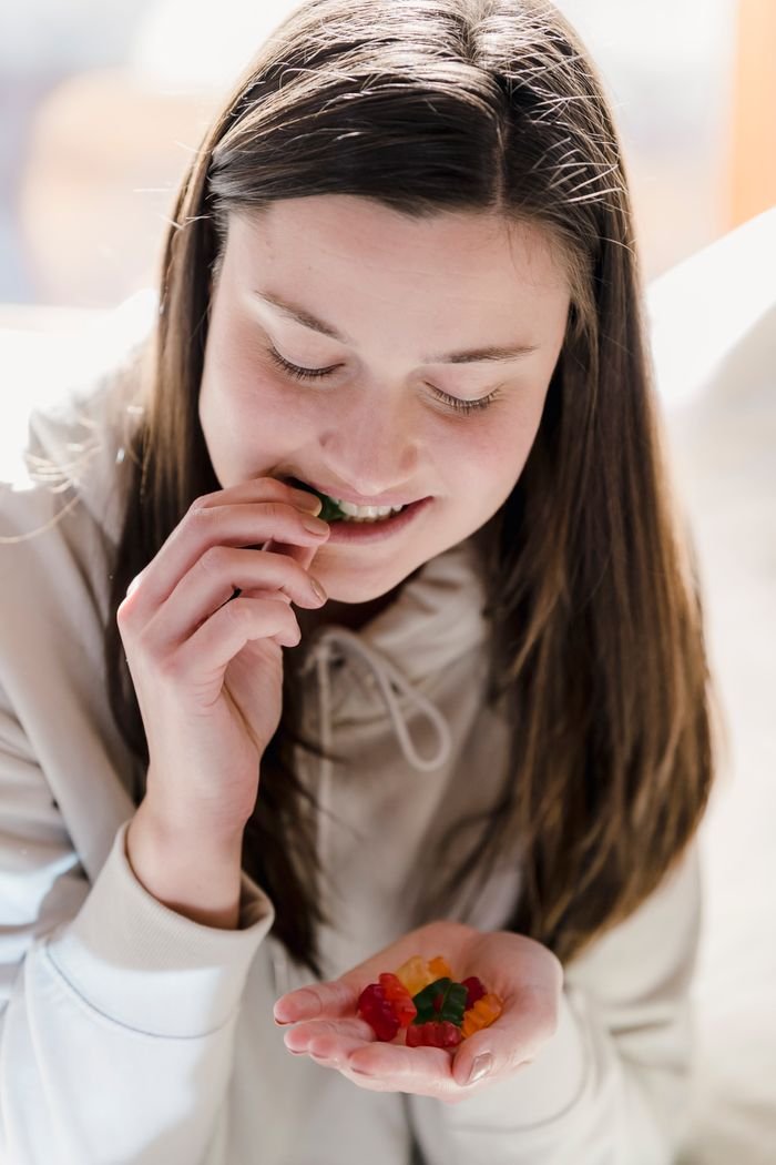 our-mission-01 Positive female millennial with long brown hair in casual clothes smiling while eating delicious sweet gum candies on sunny day at home