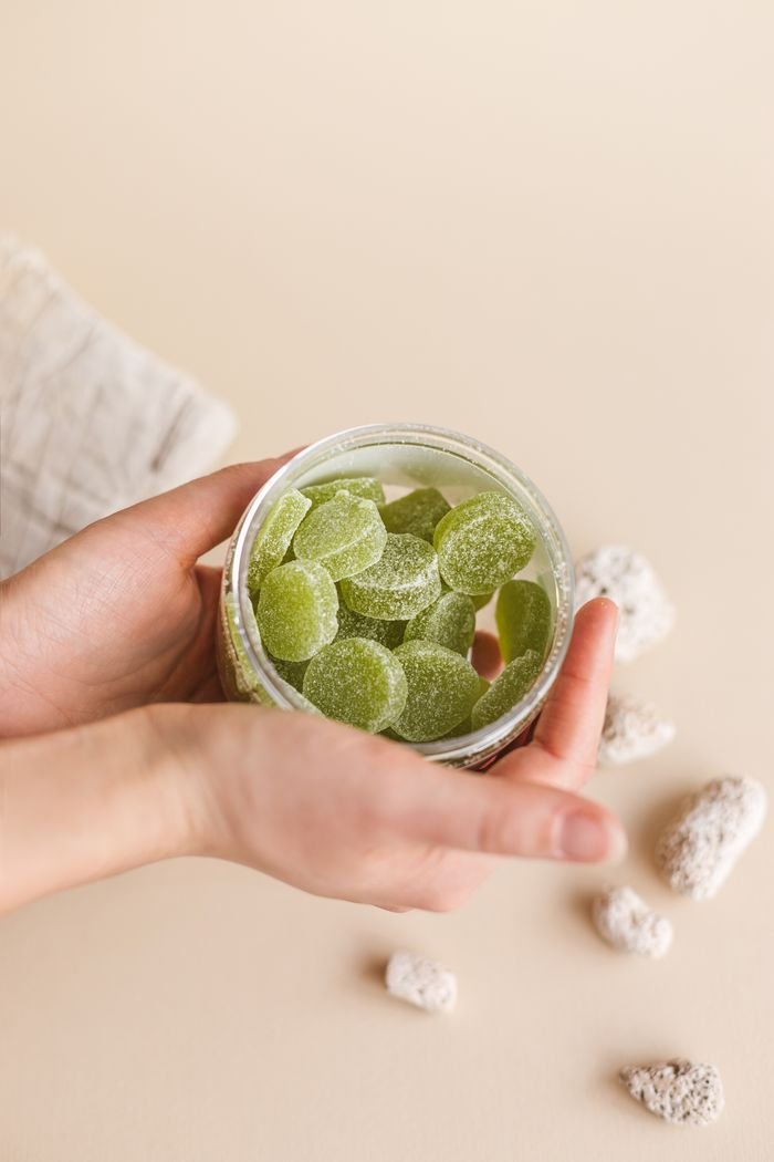 Hands holding a jar filled with green gummies against a neutral background.